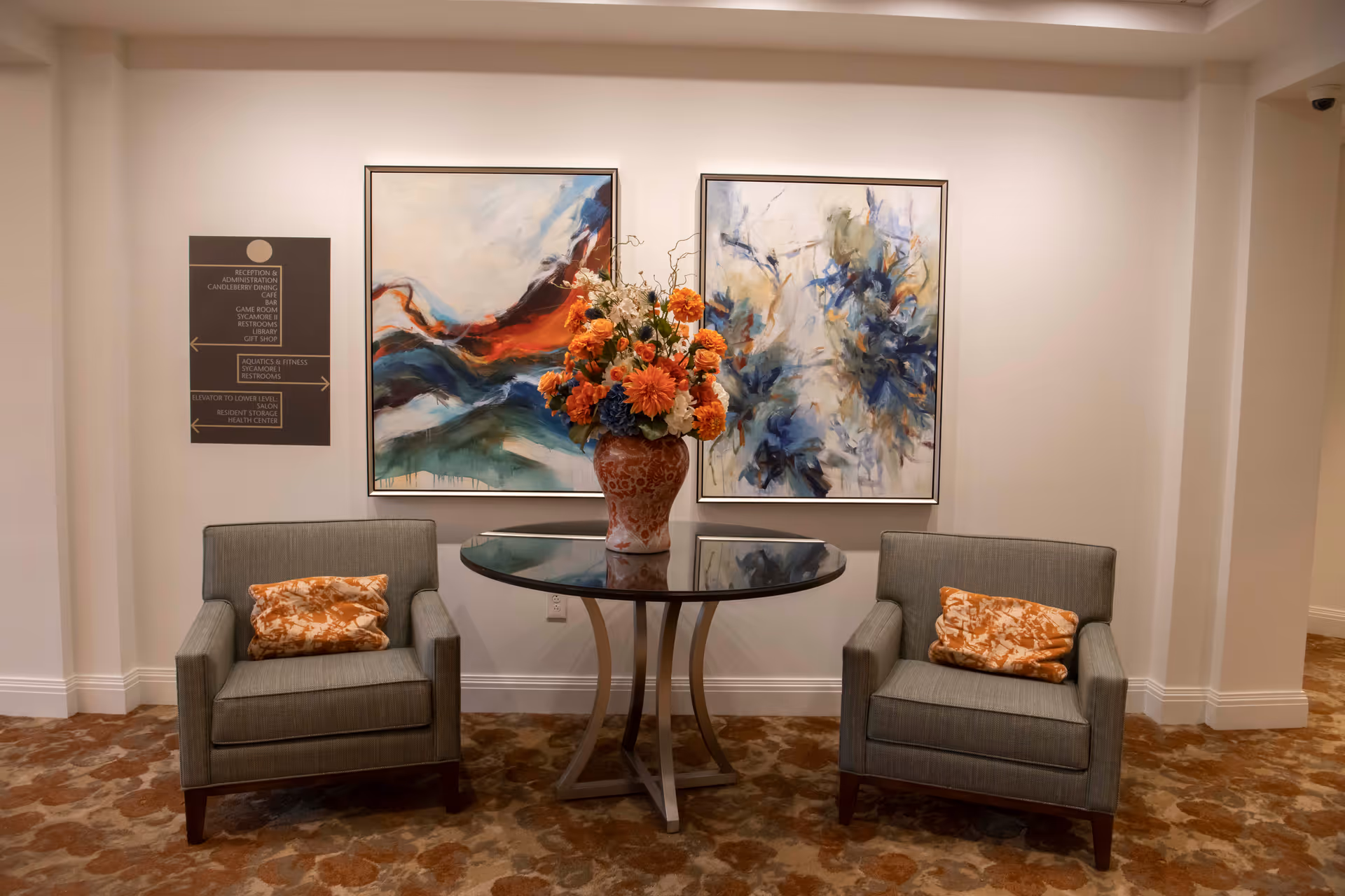 Lobby seating area with two armchairs flanking a round glass-top table topped by a large floral arrangement and abstract paintings on the wall.