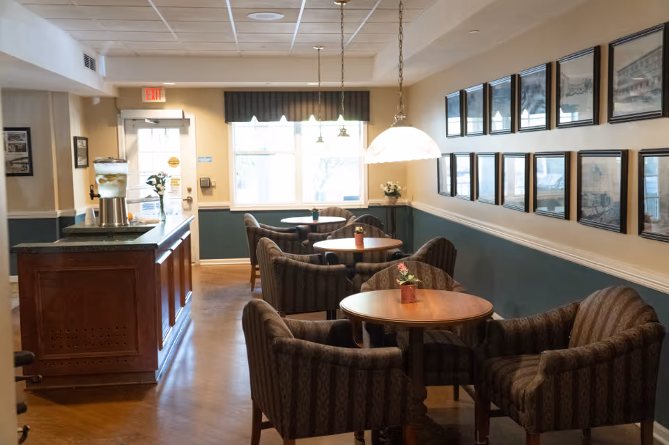 A cozy seating area with round wooden tables and cushioned armchairs arranged in pairs. The room has a wooden floor, a counter with a water dispenser, and framed pictures on the walls. Large windows let in natural light, and pendant lights hang from the ceiling.