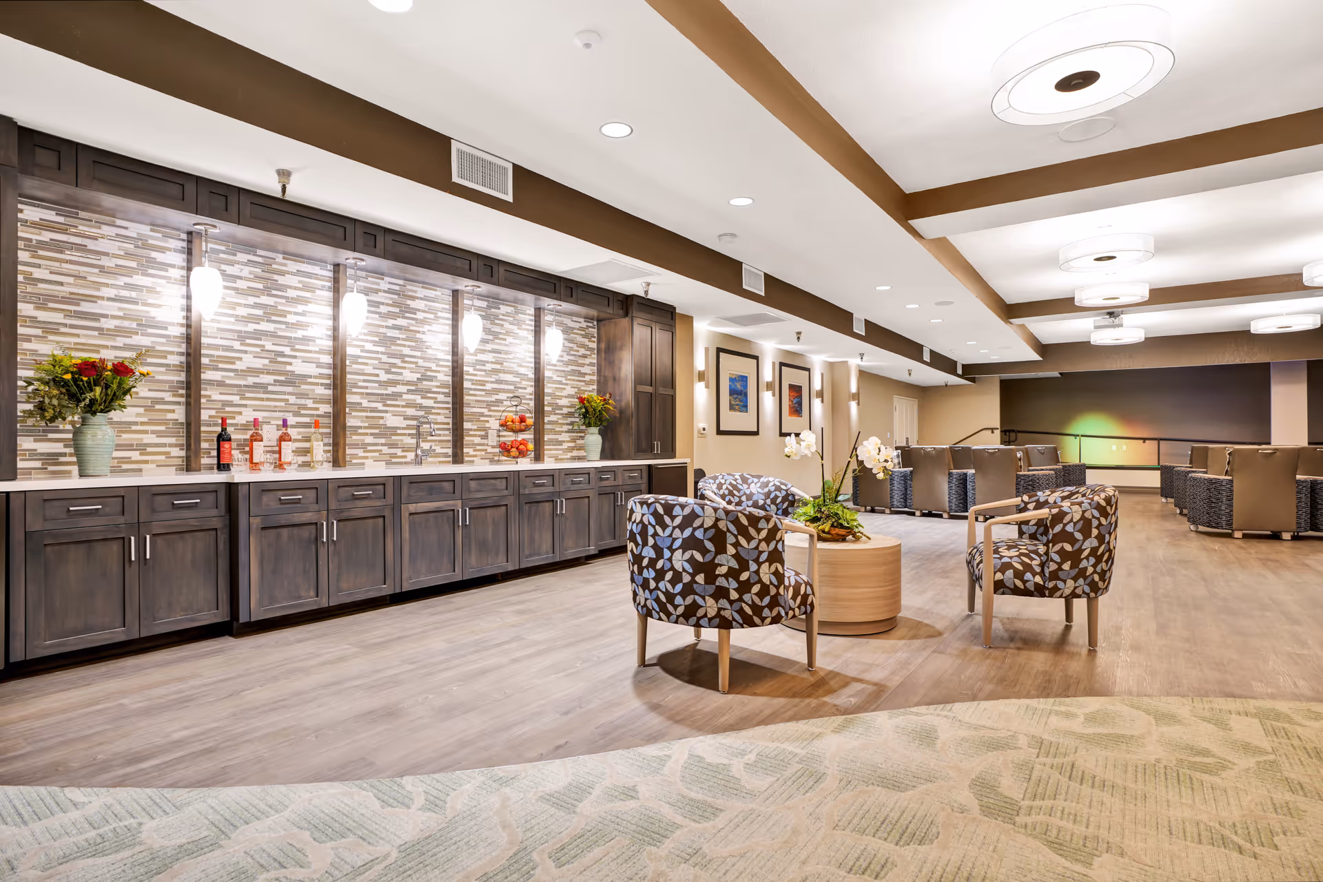 A spacious, well-lit common area with a long counter featuring dark wood cabinets and a tiled backsplash. The counter has decorative vases with flowers, bottles, and a bowl of fruit. In the foreground, there are patterned armchairs around a round wooden table with a floral arrangement. The room has recessed lighting, ceiling fixtures, and framed artwork on the walls.