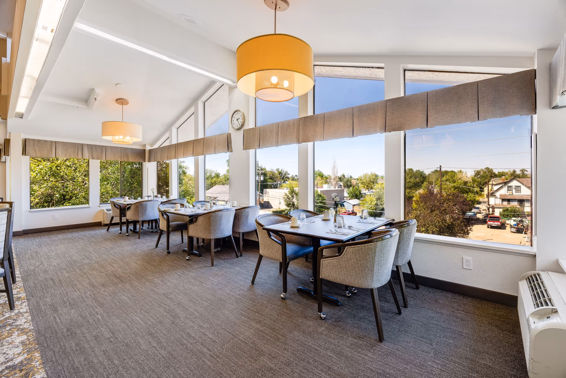 A bright dining room with large windows offering a view of trees and houses outside. The room has several tables set with glasses, plates, and utensils, surrounded by cushioned chairs. Two large pendant lights hang from the ceiling, and a clock is mounted on the wall between the windows.