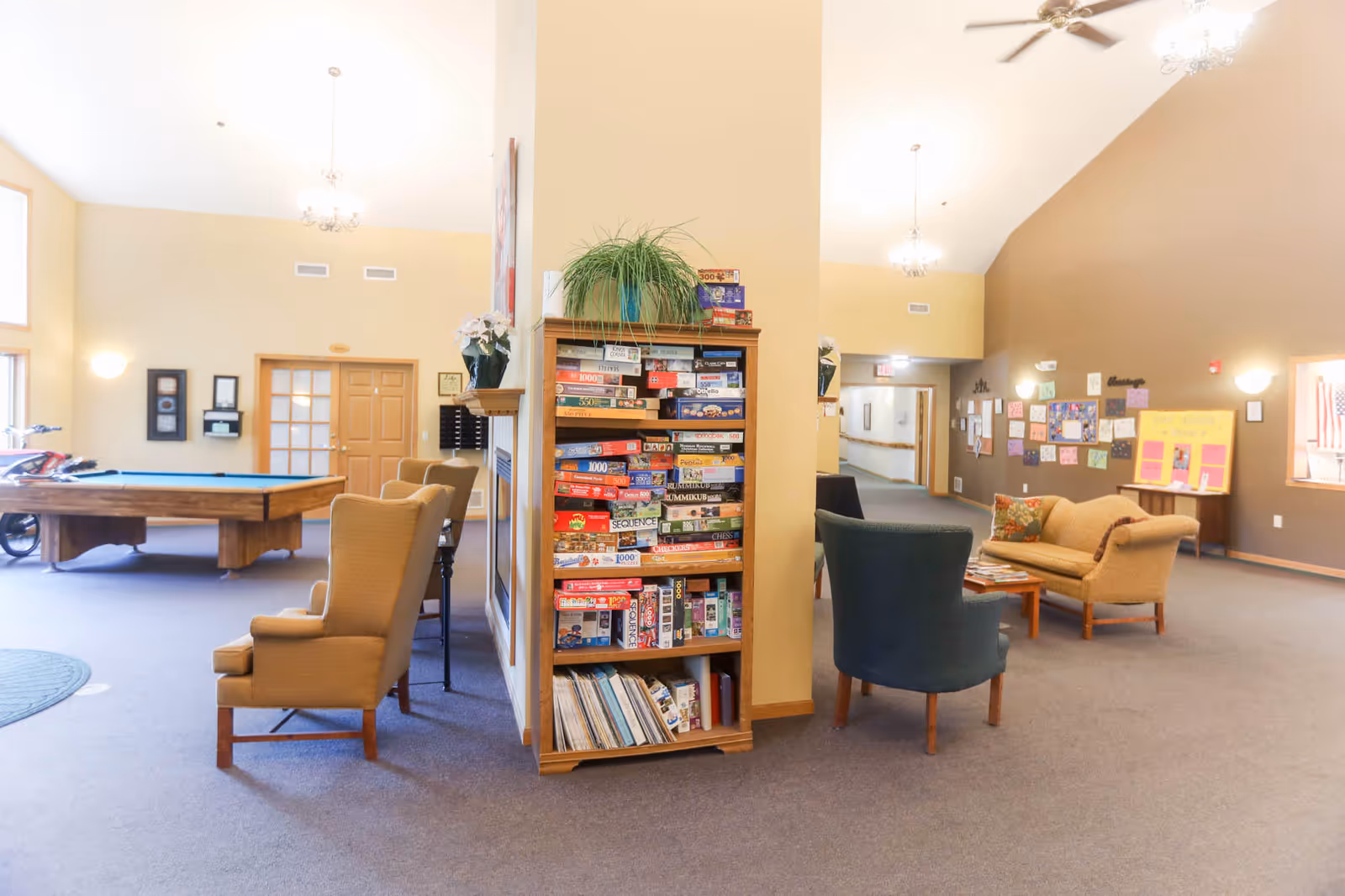 Spacious senior living common area with a central bookshelf of board games, seating areas and a pool table.