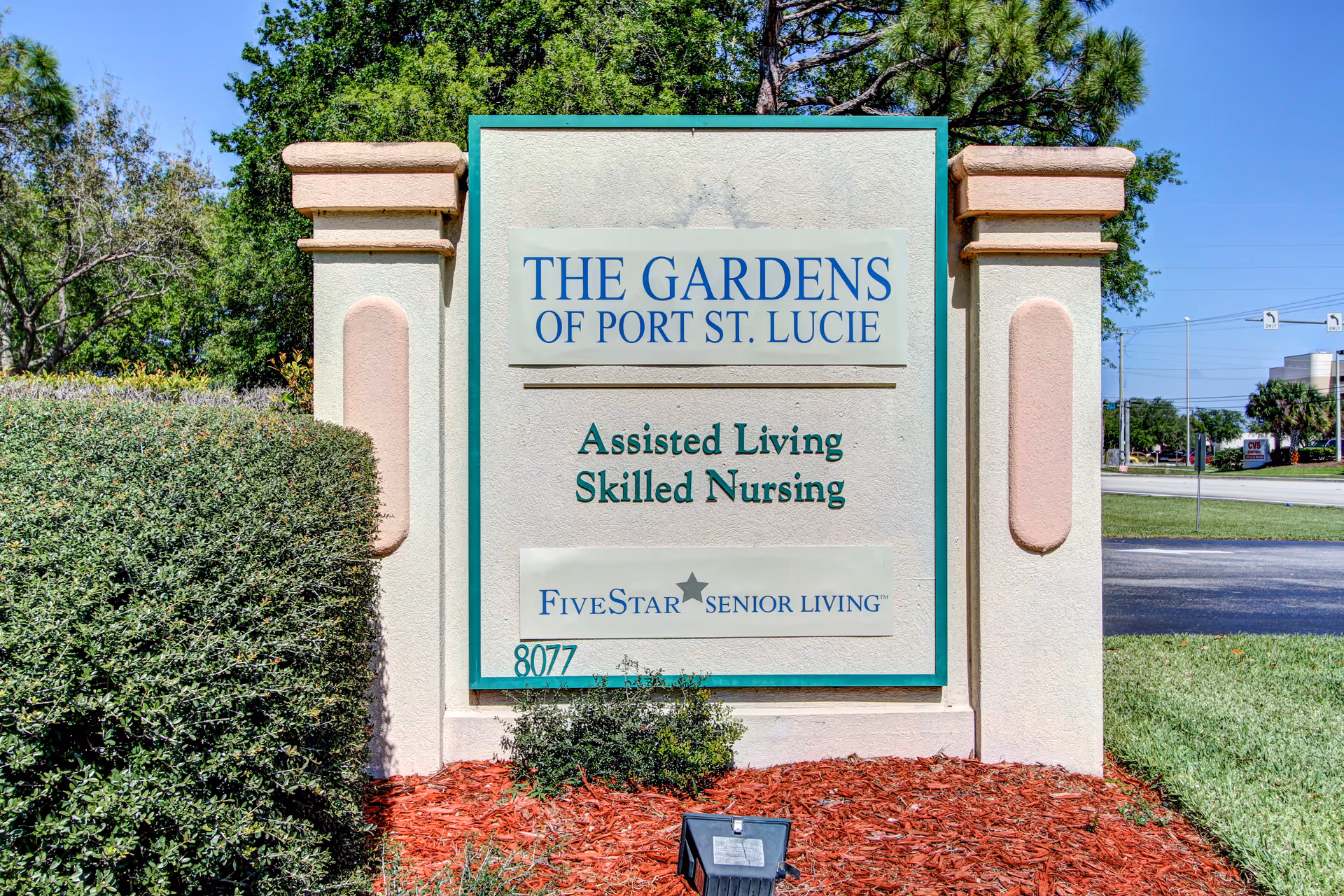 Outdoor sign for The Gardens of Port St. Lucie, an assisted living and skilled nursing facility, surrounded by landscaping with bushes, mulch, and trees in the background.