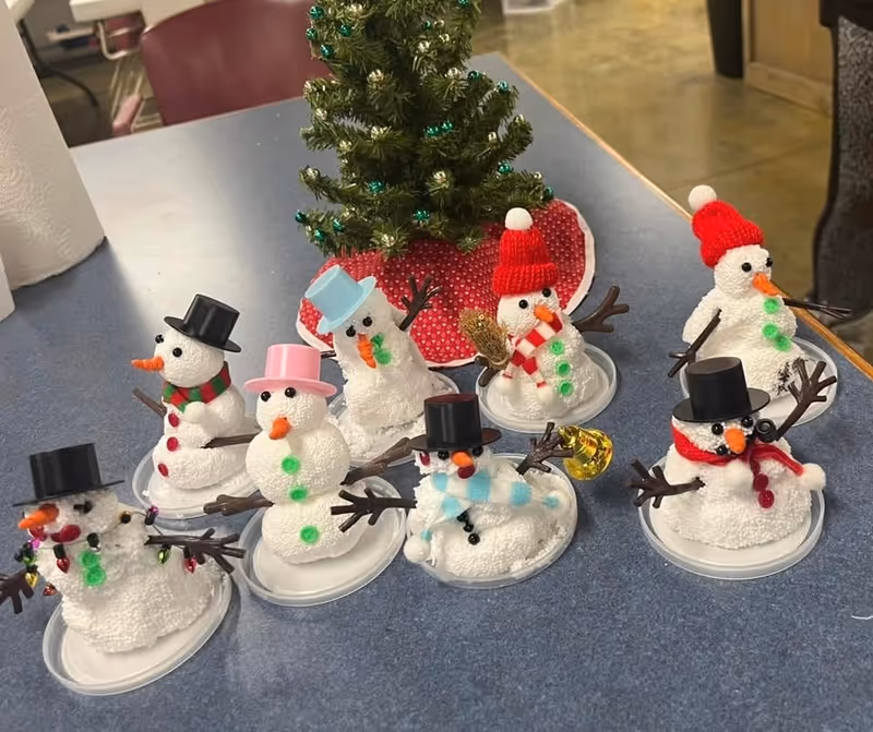 A group of eight small decorative snowmen made from white textured material, each with different colored hats, scarves, and buttons, arranged on a blue table. Behind them is a small decorated Christmas tree with green and gold ornaments on a red tree skirt. The setting appears to be indoors with chairs and a tiled floor visible in the background.
