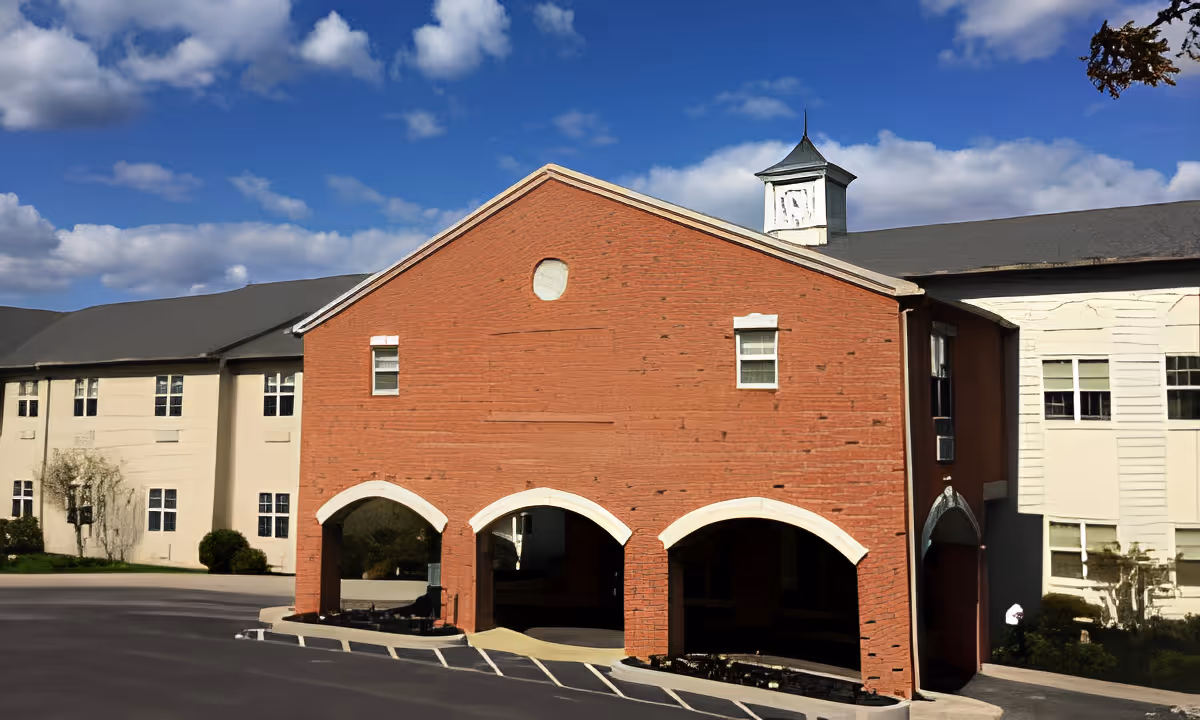 Exterior view of a senior living facility building with a red brick entrance featuring three arched openings and beige walls on either side under a partly cloudy blue sky.