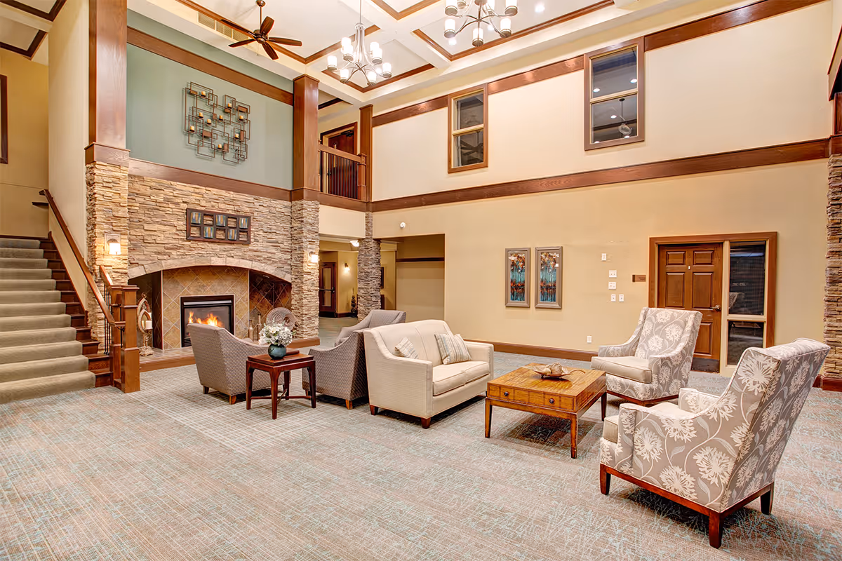 A spacious living room area in a senior living facility with a high ceiling and wooden beams. The room features a stone fireplace with a fire burning, surrounded by comfortable seating including a beige sofa, two patterned armchairs, and two gray armchairs. There is a wooden coffee table and side table with a flower vase. A staircase with carpeted steps leads to an upper floor. The walls are decorated with framed artwork and a metal wall sculpture above the fireplace.