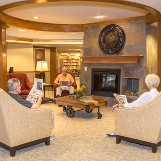 Three elderly people sitting in a cozy living room area with beige armchairs around a wooden coffee table on wheels. They are reading newspapers near a stone fireplace with a decorative clock above it. The room has warm lighting and wooden accents.