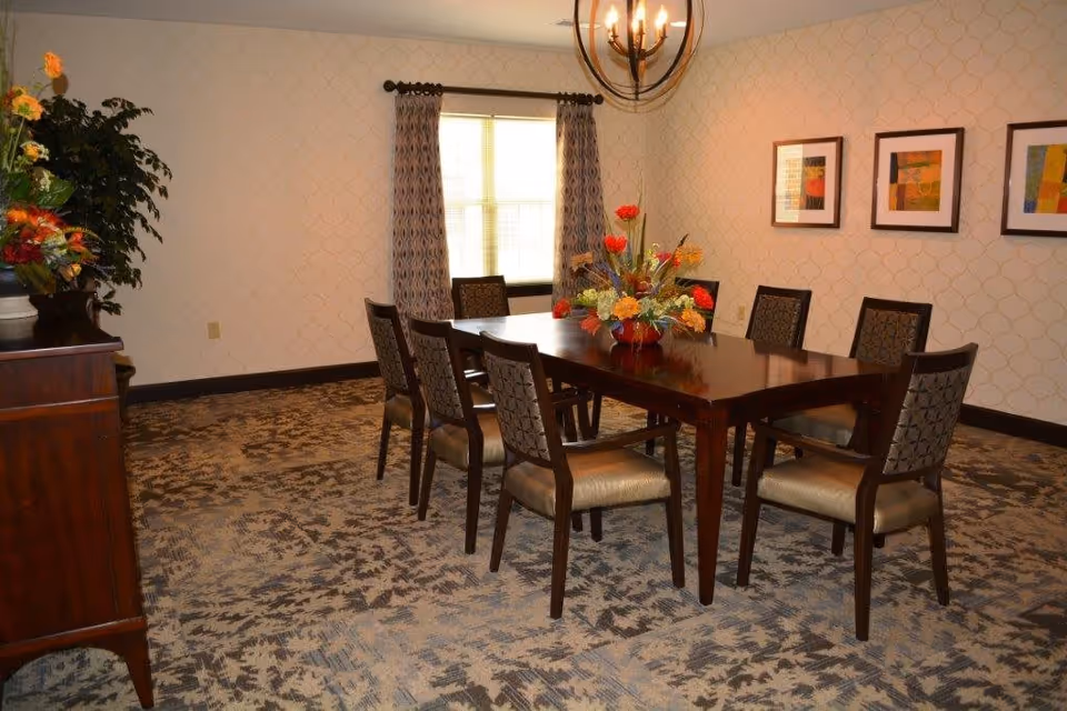 A dining room with a rectangular wooden table surrounded by eight cushioned chairs. A floral centerpiece decorates the table. The room has patterned carpet, wallpaper with a subtle geometric design, a window with curtains, three framed abstract artworks on the wall, and a chandelier hanging from the ceiling.