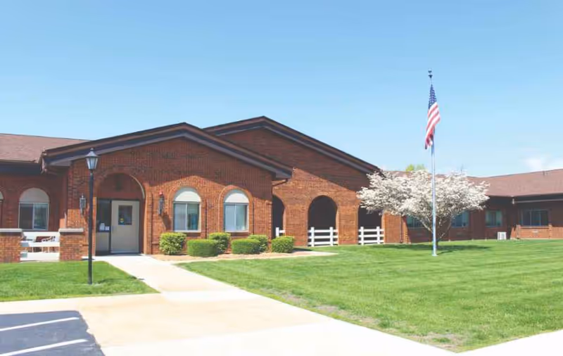 Exterior view of a single-story brick building with arched windows and doorways, a well-maintained lawn, a flowering tree, and an American flag on a flagpole under a clear blue sky.