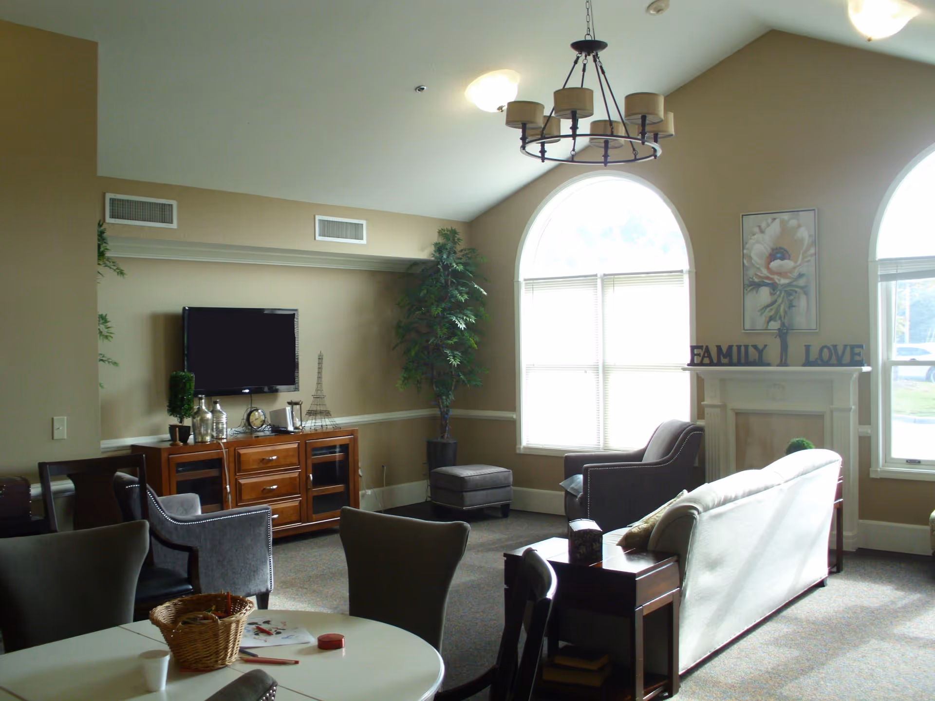 A cozy living room with beige walls and carpeted floor featuring a white couch, two gray armchairs, a wooden TV stand with a flat-screen TV mounted above it, a small ottoman, and a decorative fireplace with the words 'FAMILY' and 'LOVE' on the mantel. Large arched windows allow natural light to fill the room, and a round table with chairs and a basket of art supplies is in the foreground.