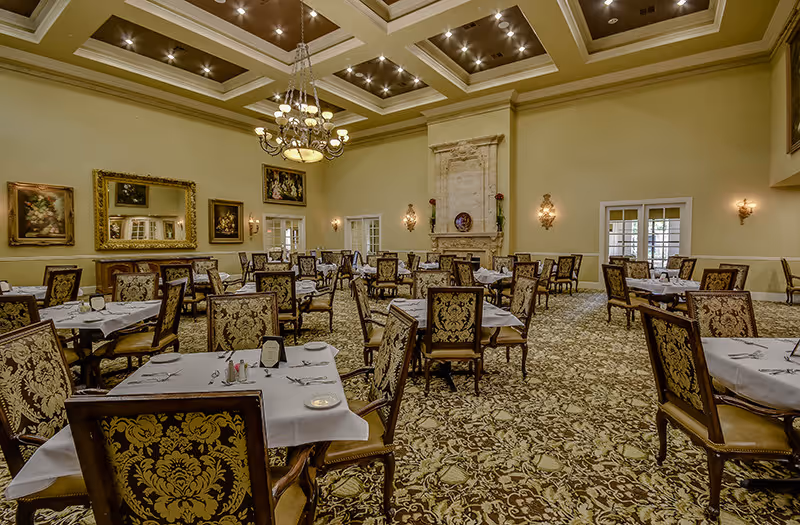 A spacious, elegant dining room with multiple tables covered in white tablecloths and set with plates, utensils, and napkins. The room features ornate upholstered chairs, a patterned carpet, a large chandelier hanging from a coffered ceiling, framed artwork on the walls, and a decorative fireplace.