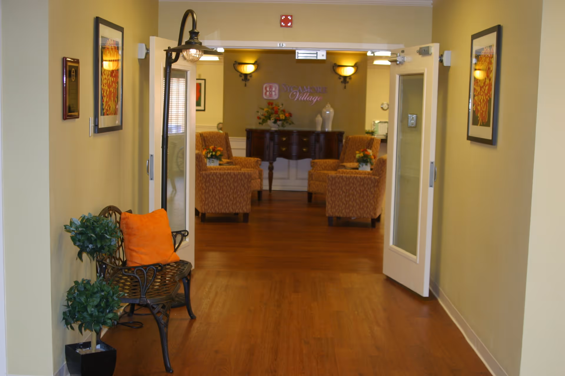View down a hallway with wooden flooring leading to a sitting area with patterned armchairs and a wooden console table with decorative vases and flowers. The walls are painted light yellow and adorned with framed artwork. A black metal bench with an orange cushion and a potted plant are positioned on the left side of the hallway. Double glass doors are open, allowing a clear view into the sitting area.