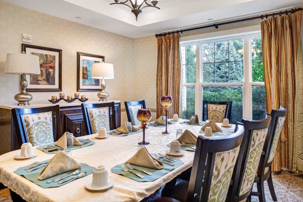 A dining room with a long table set for eight people. The table is covered with a white tablecloth and has beige napkins folded on green placemats, white cups, glasses, and silverware. Two decorative goblets are placed in the center of the table. The room has patterned wallpaper, two framed pictures on the wall, two table lamps on a sideboard, and large windows with striped curtains showing greenery outside.