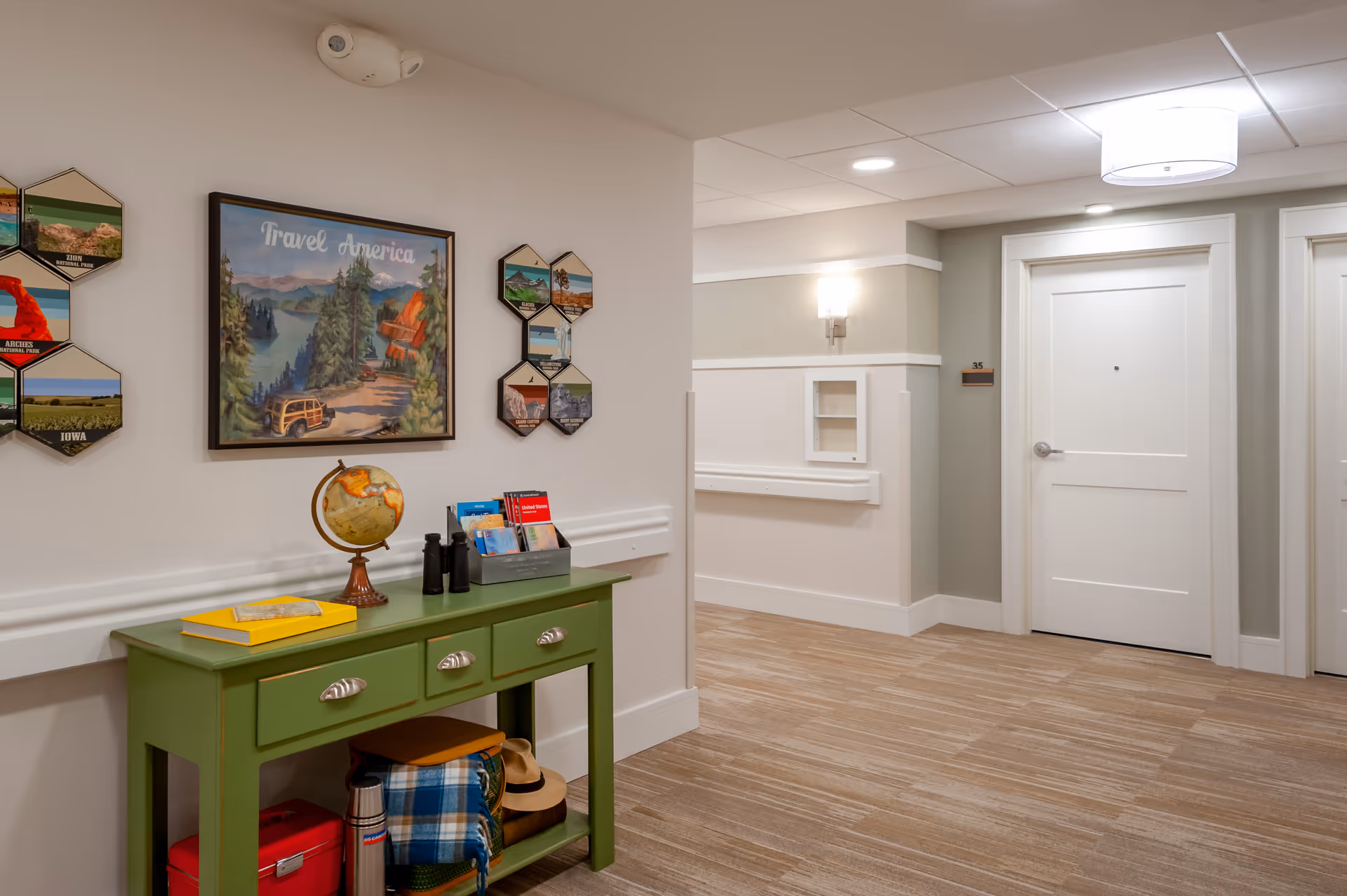 A well-lit hallway in a senior living facility with beige walls and carpeted floor. There is a green console table against the wall with a globe, binoculars, books, and a yellow box on top. Below the table are a red cooler, a thermos, a plaid blanket, and a hat. On the wall above the table are travel-themed decorations including a framed picture titled 'Travel America' and several hexagonal photo displays. The hallway has white doors and a modern ceiling light fixture.