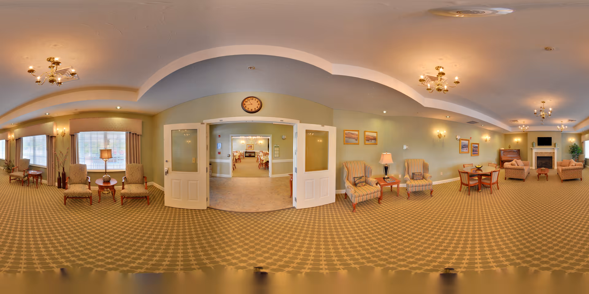 A spacious, well-lit common area in a senior living facility with patterned carpet, multiple seating arrangements including armchairs and small tables, wall-mounted lamps, framed artwork, and a fireplace. Double doors lead to an adjoining dining room with tables and chairs. The walls are painted light green and the ceiling has recessed lighting and chandeliers.