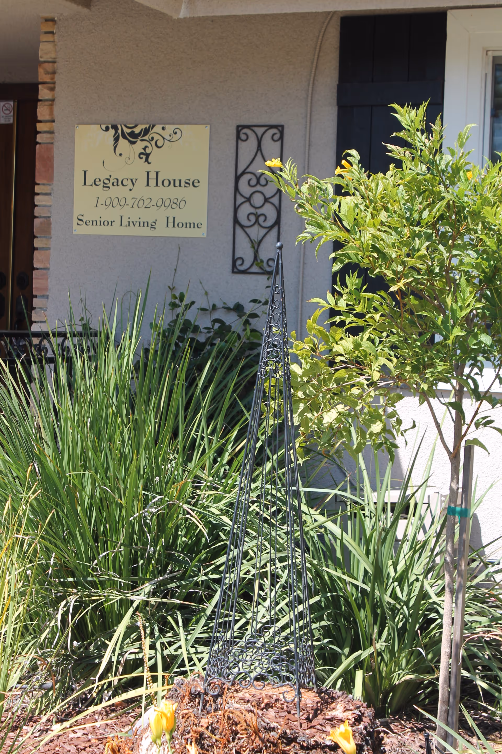 Front garden of Legacy House senior living home with a wall sign, ornamental metal trellis and lush green plants.