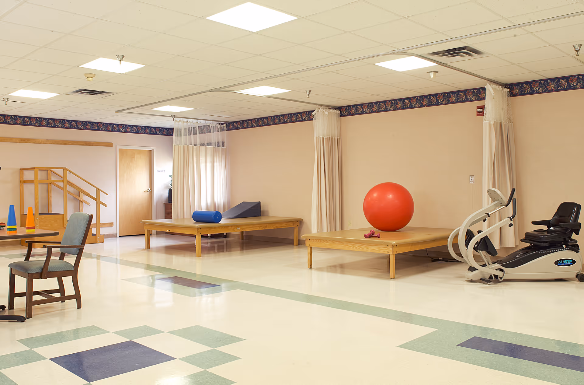 A spacious physical therapy or rehabilitation room with exercise equipment including a recumbent exercise bike, two wooden therapy platforms with exercise props such as a large red exercise ball, a blue foam roller, and a wedge cushion. There are privacy curtains hanging from the ceiling, a small table with chairs, and a wooden staircase for mobility exercises. The room has a tiled floor with a geometric pattern and beige walls with a floral border near the ceiling.