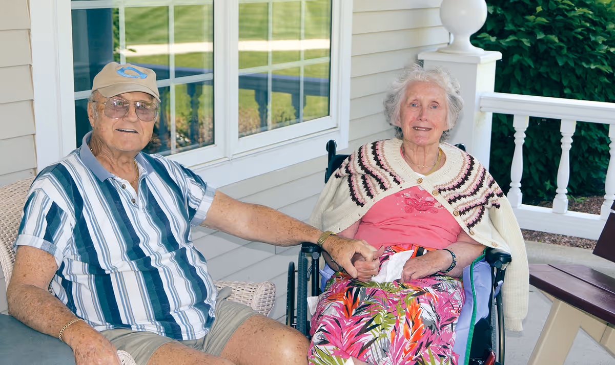 An elderly man and woman sitting together on a porch. The man is seated on a wicker chair wearing a striped shirt, shorts, glasses, and a cap, holding hands with the woman who is in a wheelchair wearing a pink top, colorful skirt, and a knitted shawl. They are outside a building with beige siding and white trim, with greenery visible in the background.