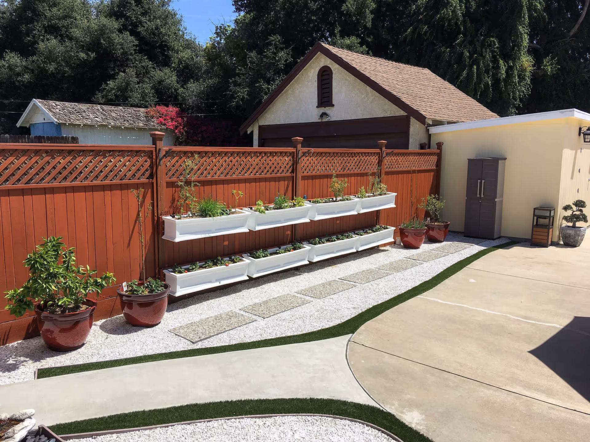 Outdoor garden area with a red wooden fence featuring white planter boxes filled with various plants. There are several large red pots with plants on the ground, a small shed or garage with a brown roof in the background, and a paved walkway with stepping stones and artificial grass borders.