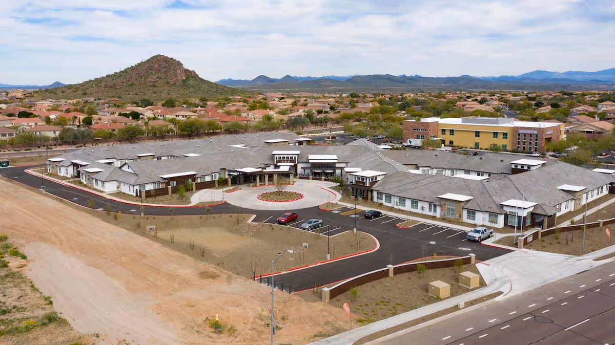 Aerial view of Spring Gardens Senior Living Peoria facility showing a large single-story building with a circular driveway and parking area in front. The building is surrounded by a residential neighborhood with many houses and a mountain in the background under a partly cloudy sky.