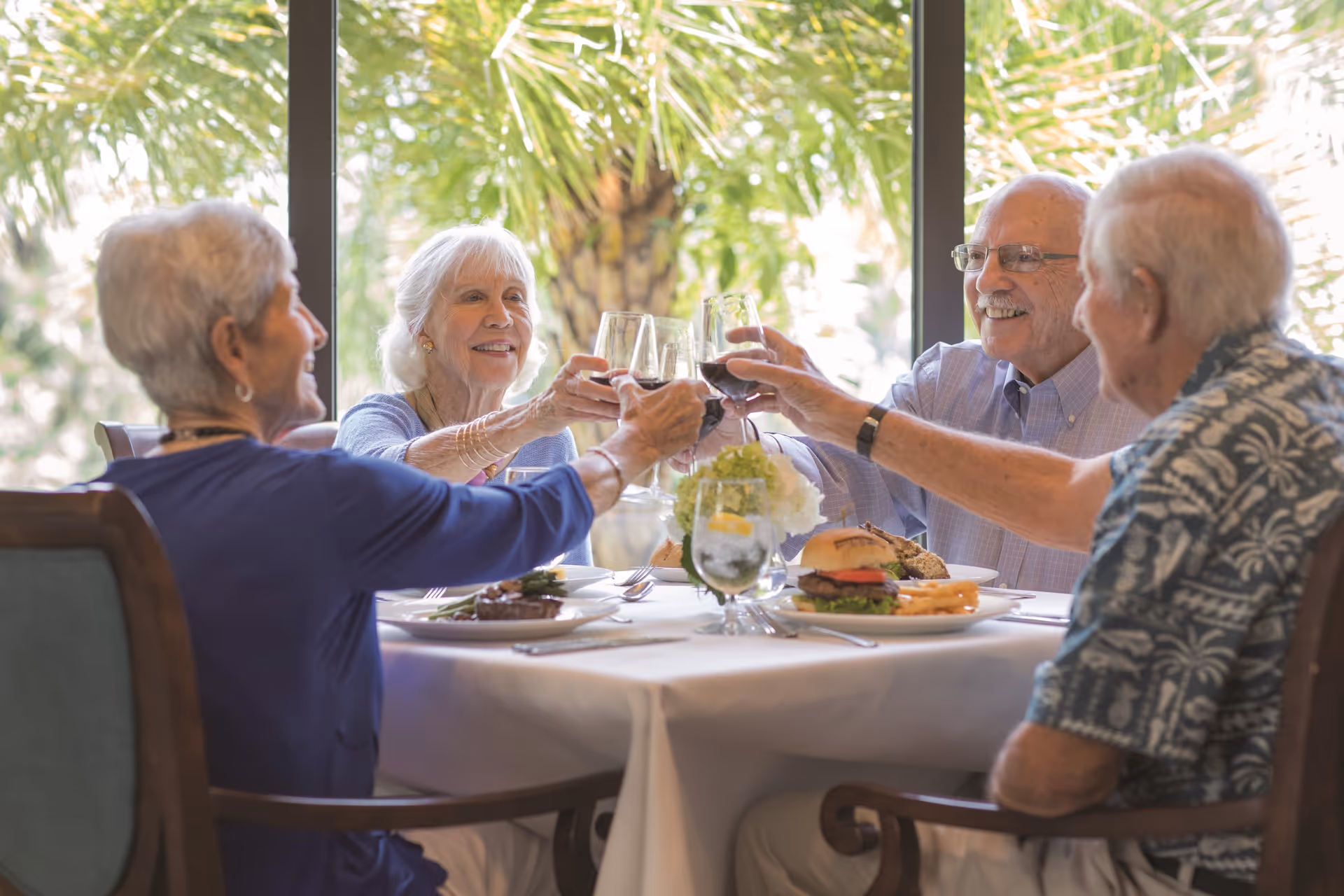 Four elderly people sitting around a dining table, smiling and toasting with glasses of red wine. The table is set with plates of food including a burger and fries, and there is a large window with greenery visible outside.