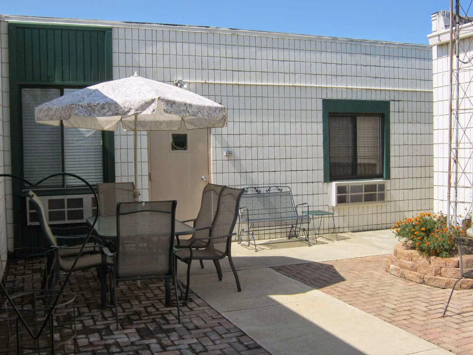 Outdoor patio area with a table and six chairs under a large umbrella, a metal bench, two windows with air conditioning units, and a small flower bed with orange flowers against a white building wall.