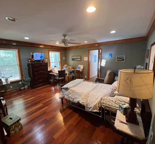 A cozy assisted living bedroom with wooden floors and green walls. The room features a bed with floral bedding, a bedside table with a lamp, a recliner chair, a dresser with a TV on top, and a small desk with a chair near two windows. There are framed pictures on the walls and a ceiling fan with lights.