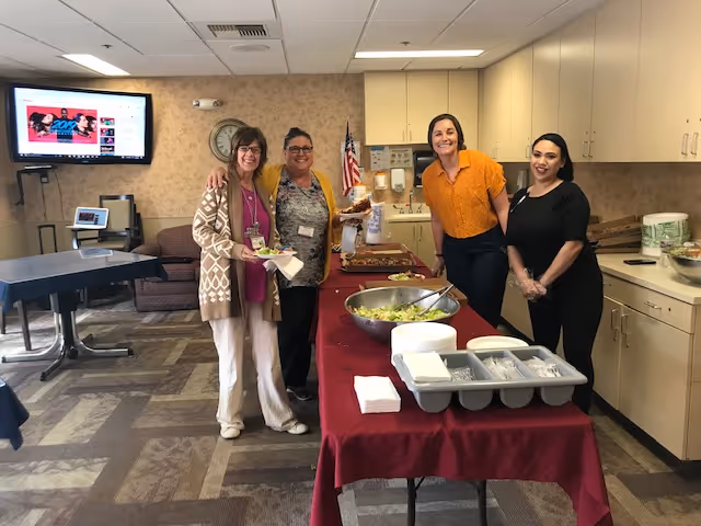 Four women standing behind a table with food in a room with beige walls and cabinets. The table is covered with a red tablecloth and has a large bowl of salad, plates, and utensils. There is a television mounted on the wall, a clock, and an American flag in the background.
