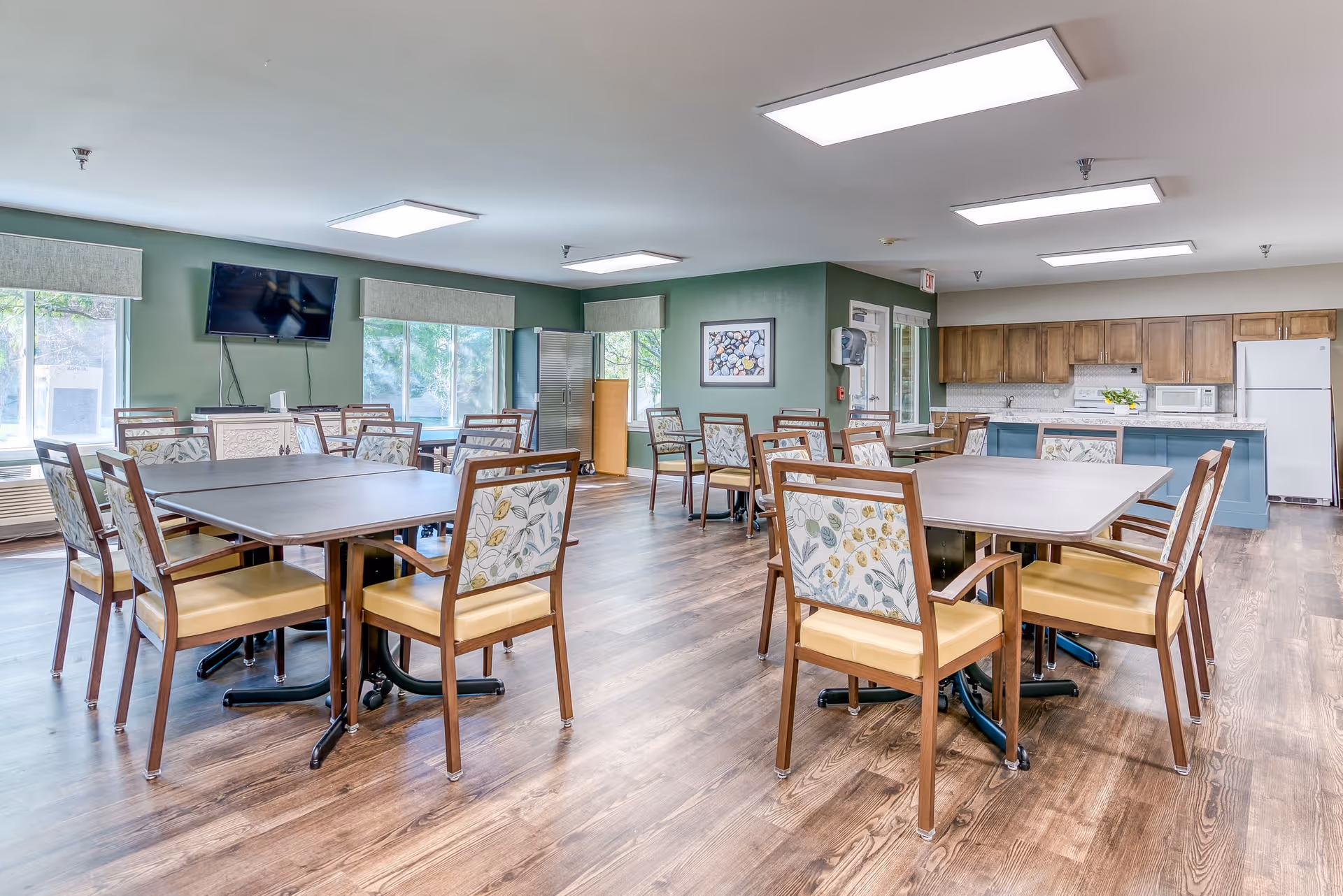 A bright and spacious dining area in a senior living facility with multiple wooden tables and chairs featuring floral upholstery. The room has large windows letting in natural light, a mounted flat-screen TV on a green wall, and a kitchen area with wooden cabinets, a refrigerator, microwave, and sink in the background.