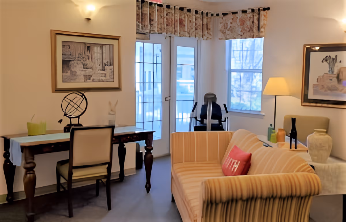 A cozy living room area with a striped sofa featuring a red pillow, a wooden desk with a chair, decorative items including a globe and vases, a floor lamp, framed artwork on the walls, and large windows with sheer curtains letting in natural light.