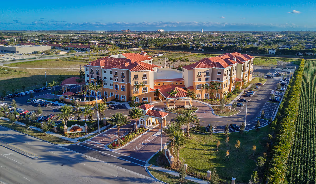 Aerial view of The Palace Gardens - Assisted Living Retirement Community, showing a large, multi-story building with terracotta roofs, surrounded by palm trees, parking lots, and landscaped grounds under a clear blue sky.