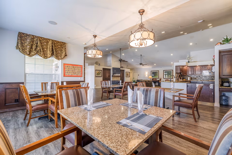 A spacious and well-lit dining area in a senior living facility with multiple tables and chairs arranged neatly. The tables have granite tops with placemats and folded napkins. The room features wooden flooring, decorative hanging light fixtures, large windows with patterned valances, and a kitchen area with dark wood cabinets in the background.