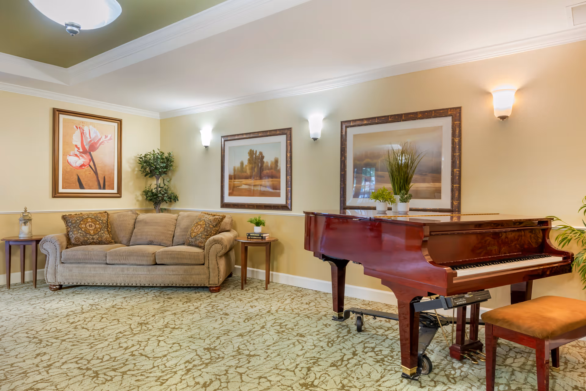A cozy lounge with a beige sofa, framed artwork, plants and a polished grand piano on patterned carpet.