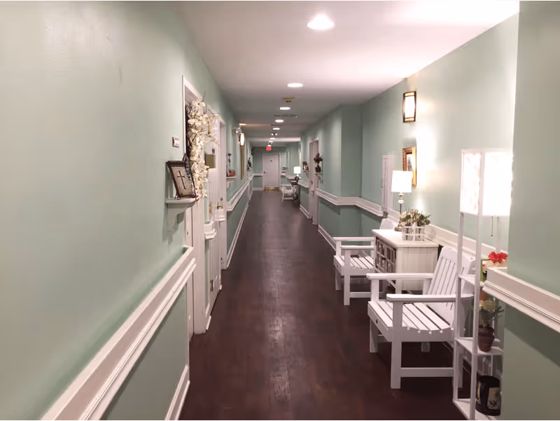 A long, well-lit hallway in a senior living facility with light green walls and dark wooden flooring. The hallway is decorated with white wooden chairs, small tables with lamps, and various plants and decorative items along the walls. Several doors line the hallway, and ceiling lights illuminate the space.