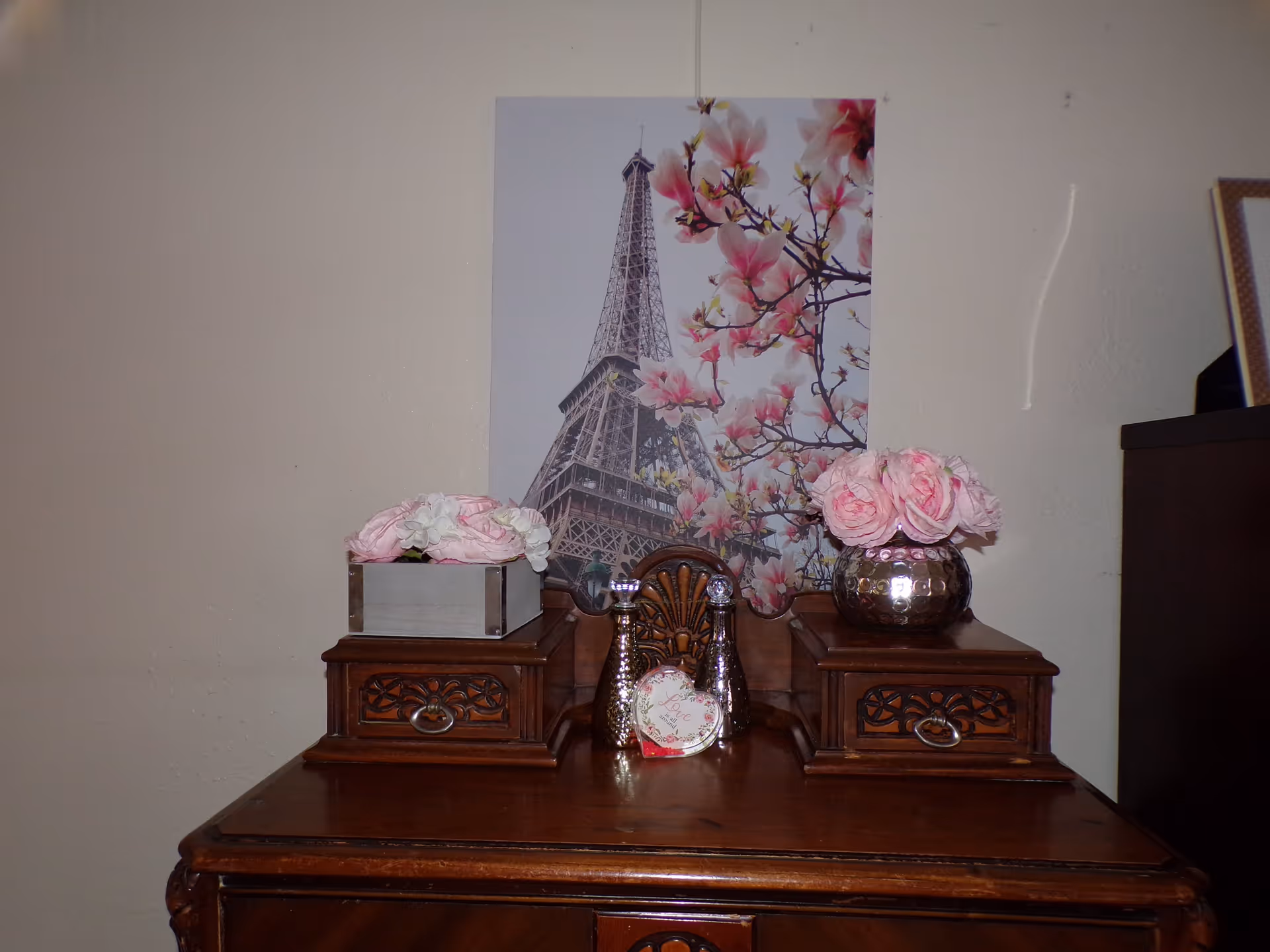 Wooden dresser topped with floral arrangements and decorative items beneath an Eiffel Tower poster on the wall.