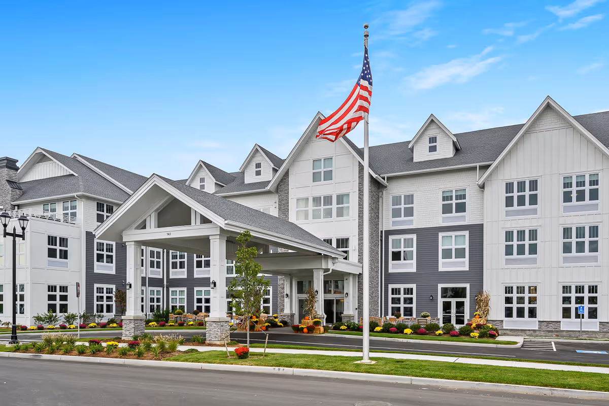 Front exterior view of a large, modern senior living facility building with white and gray siding, multiple windows, a covered entrance, landscaped flower beds, and an American flag on a flagpole in front.