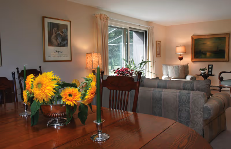 A cozy living room and dining area with a wooden dining table adorned with a centerpiece of bright sunflowers and two green candles in silver holders. The living room features patterned sofas, a large window with beige curtains, a floor lamp, framed artwork on the walls, and a small plant on a side table.