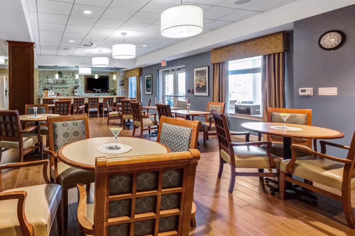 A spacious dining room with multiple round tables and wooden chairs with cushioned seats and backs. The room has wooden flooring, large windows with curtains, and modern ceiling lights. A clock is mounted on the wall, and a television is visible in the background above a stone fireplace.