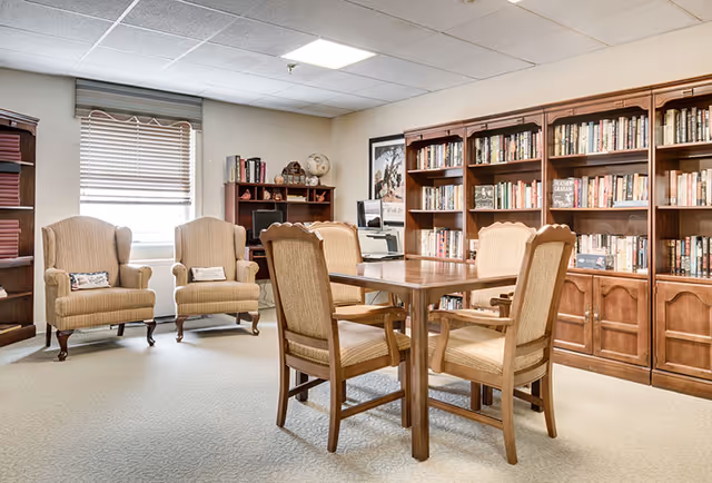 A cozy reading or meeting room with a wooden table surrounded by four upholstered chairs in the center. Behind the table, there are large wooden bookshelves filled with books. To the left, two cushioned armchairs are placed near a window with blinds partially closed. The room has a neutral color palette with beige carpet and walls, and a ceiling with recessed lighting.