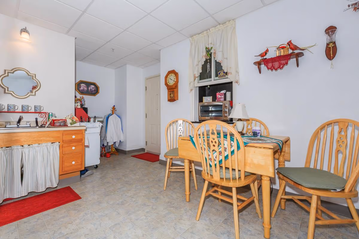 A cozy dining area in an assisted living facility featuring a wooden dining table with four matching chairs. The table has a decorative table runner and a small lamp. On the wall above the table, there is a shelf with red cardinal bird decorations and a small clock. To the left, there is a wooden cabinet with a sink and several mugs on the counter. The floor is tiled, and the walls are painted white with some framed pictures and a wall clock. A window with a lace curtain is visible behind the table.