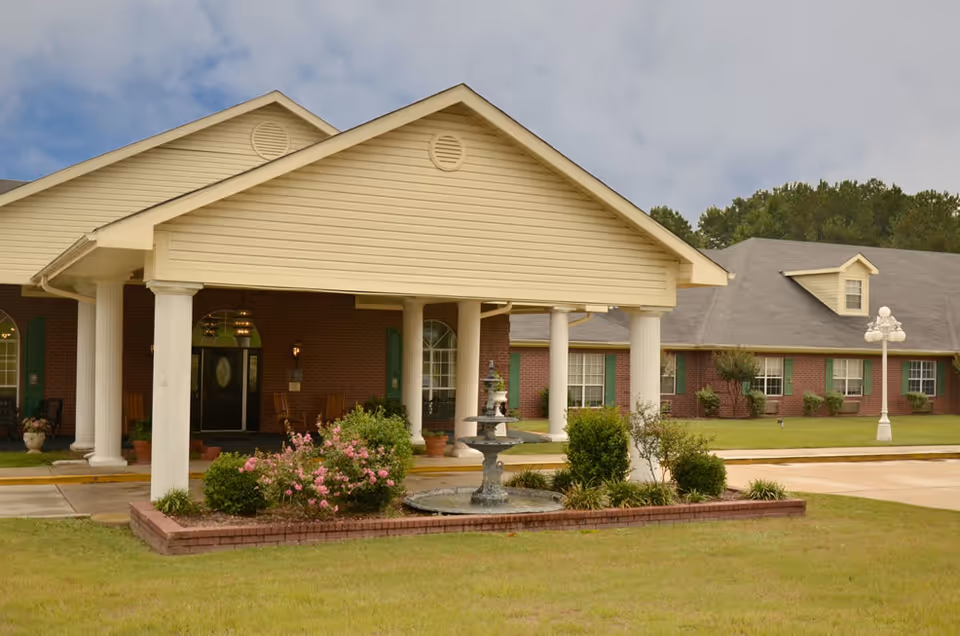 Front entrance of a brick senior care building with a covered portico supported by white columns, a central fountain and landscaped flower beds.
