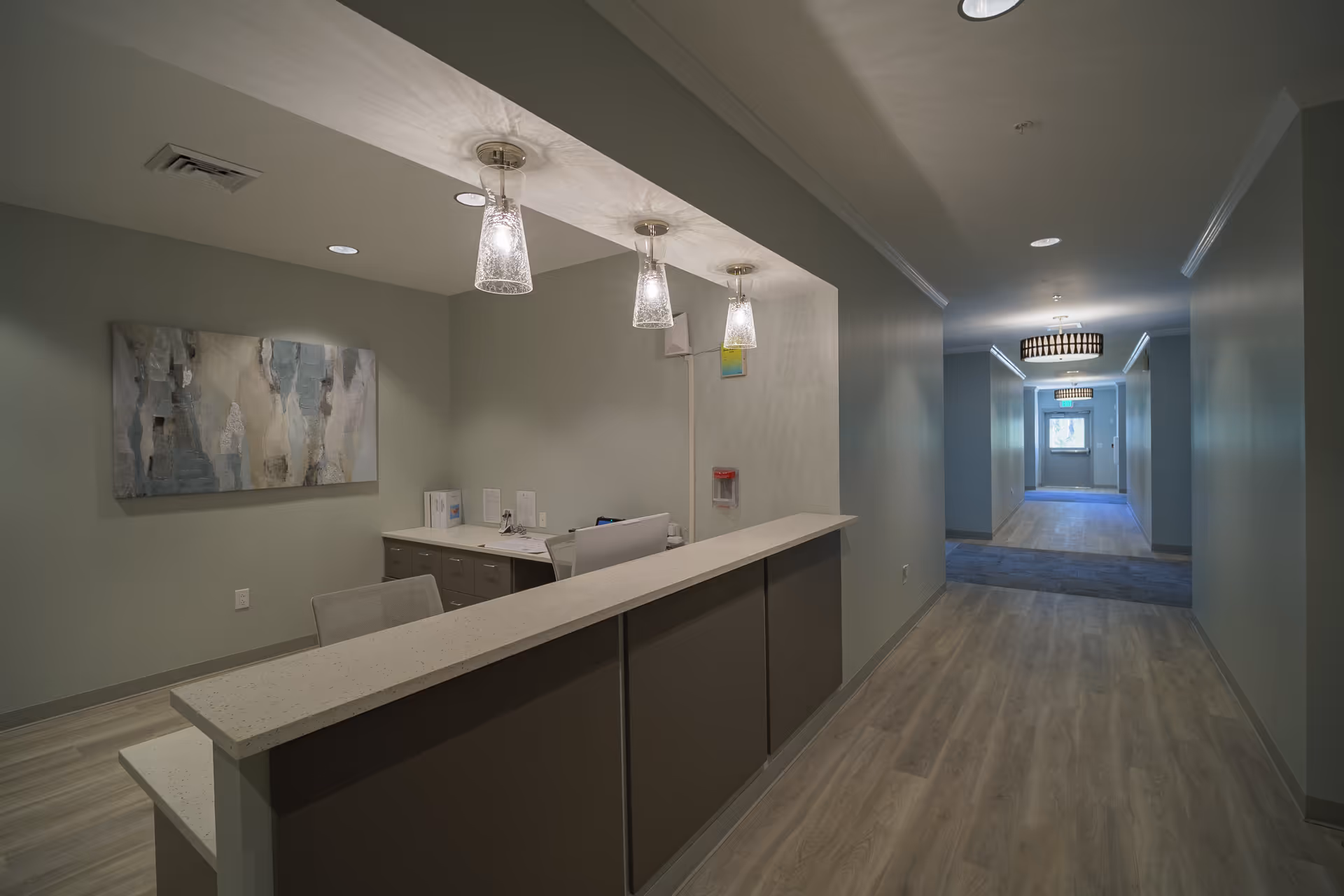 A modern reception desk area in a hallway with three pendant lights hanging above the counter. The desk has a computer and some documents on it. The hallway has light wood flooring, soft green walls, and decorative ceiling lights further down the corridor.