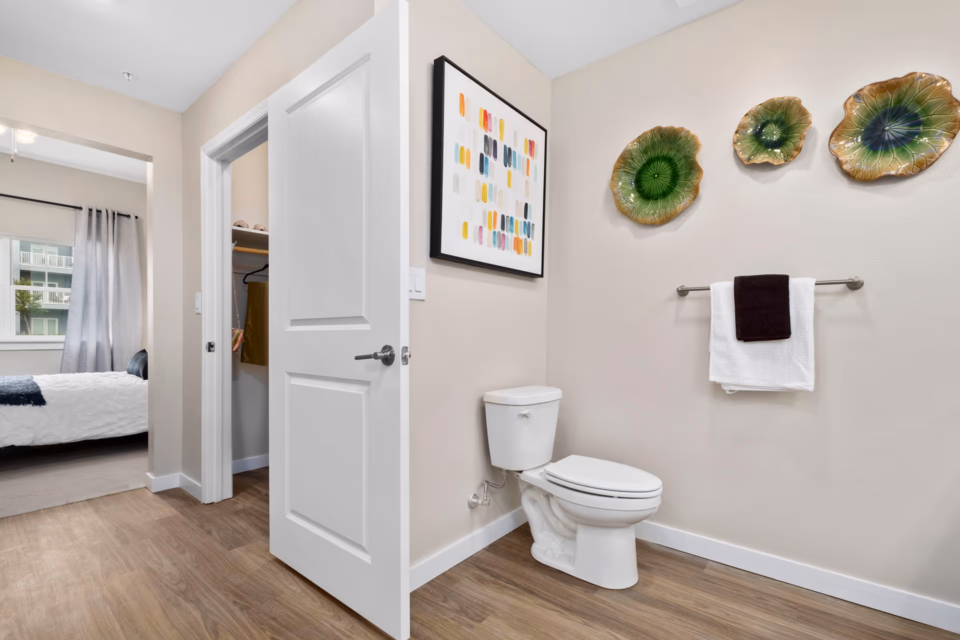 Bathroom with a white toilet, towel rack and decorative wall plates, the open door reveals a bedroom.