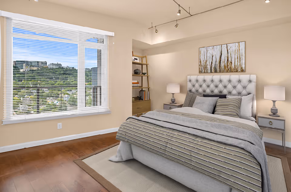 Bright bedroom with a tufted headboard, striped bedding, bedside tables and lamps, shelving, and a large window overlooking a green hillside.
