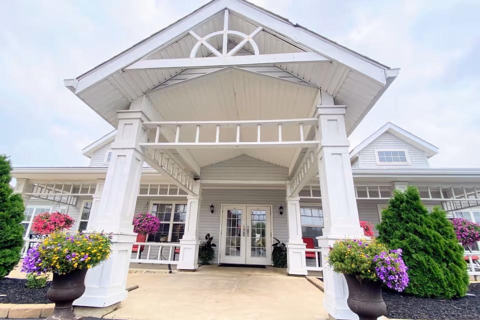 Front entrance of a white building with a covered porch supported by large white columns. There are colorful flower pots with purple, yellow, and pink flowers on either side of the entrance, along with green shrubs. The building has large windows and a double glass door under the porch roof.