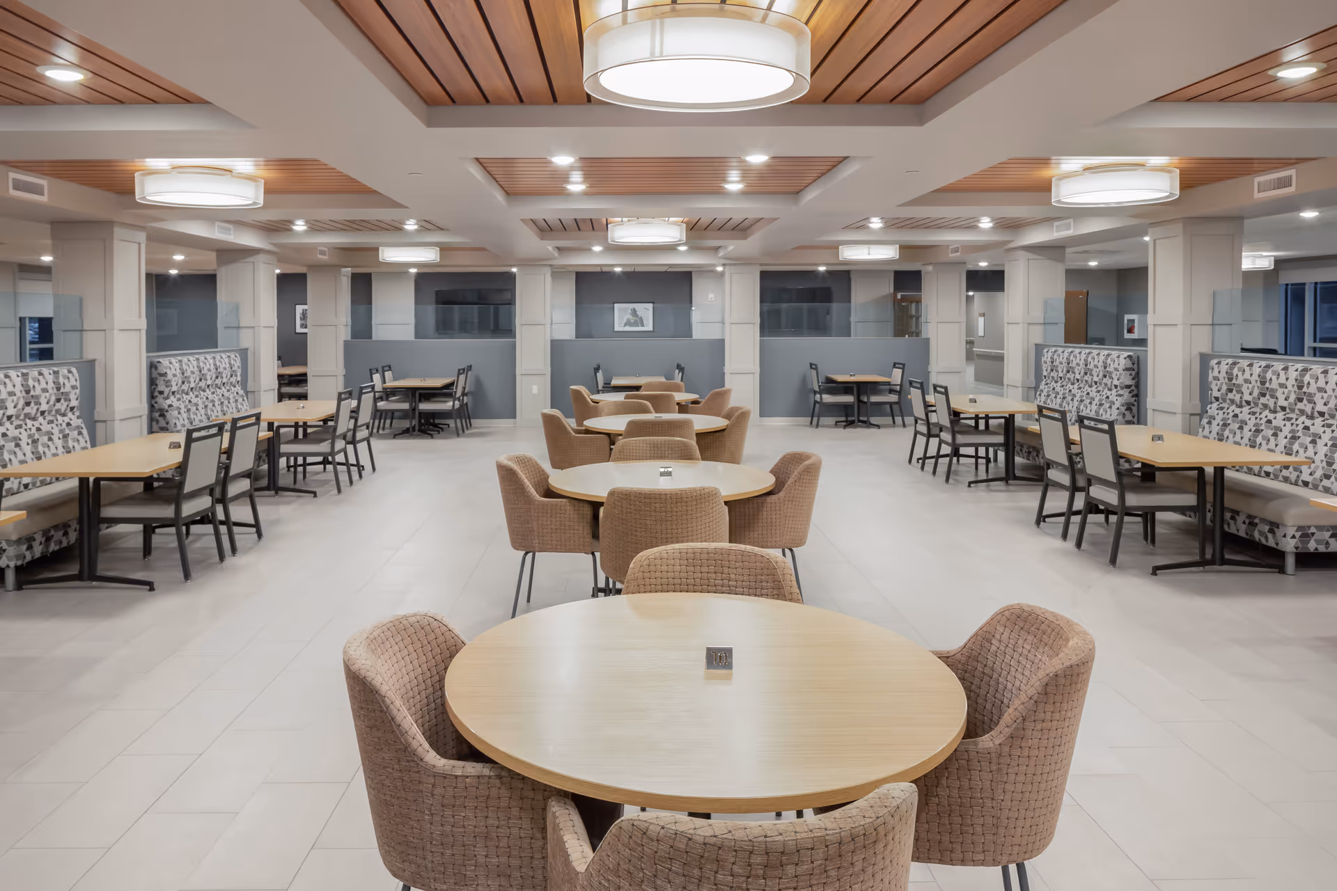 A spacious, modern dining room with round and rectangular wooden tables surrounded by cushioned chairs and booth seating. The ceiling features wooden paneling and multiple circular light fixtures, creating a warm and inviting atmosphere.