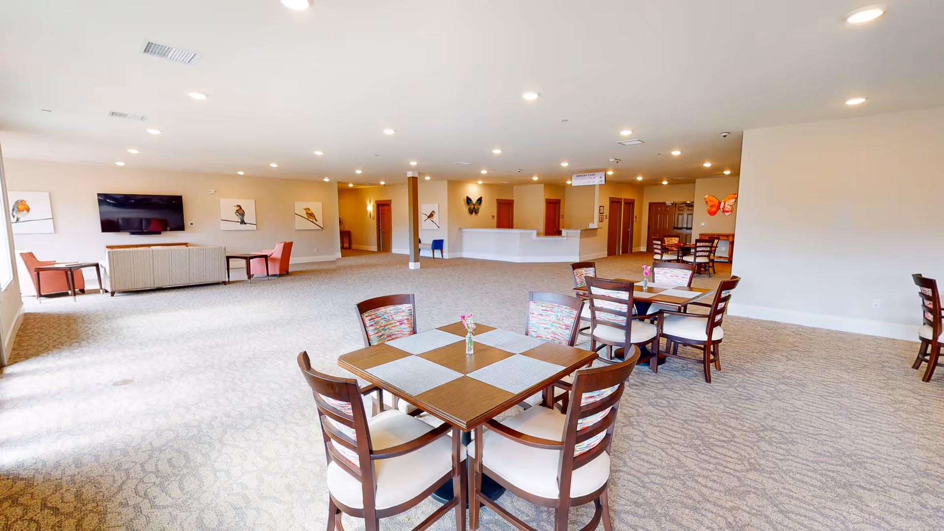 A spacious senior living facility common area with multiple wooden tables and chairs arranged for dining or socializing. The room has beige carpet and walls, with several bird-themed paintings and butterfly decorations on the walls. There is a seating area with a sofa and armchairs near a wall-mounted flat-screen TV. The area is well-lit with recessed ceiling lights.