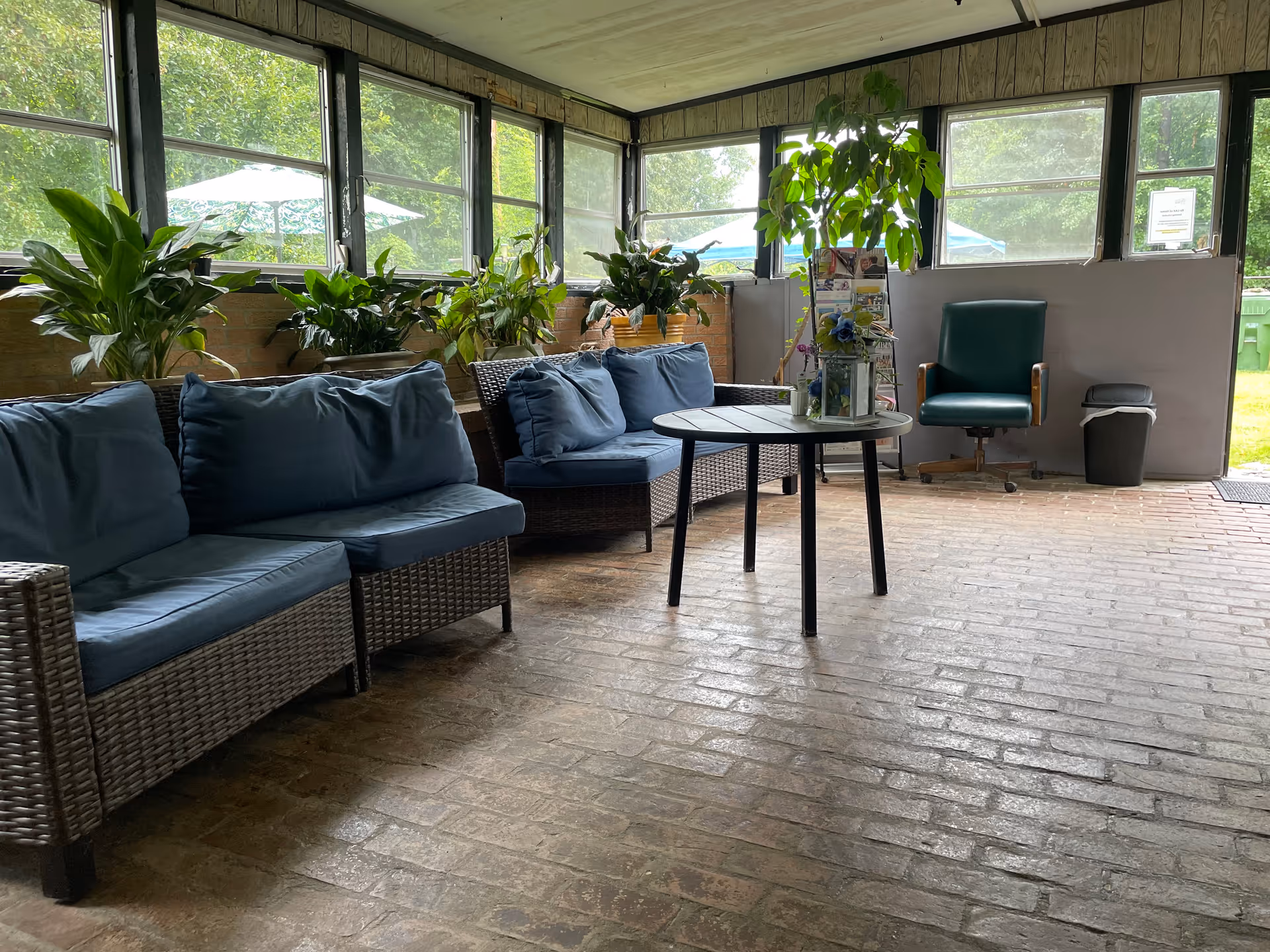 A bright indoor seating area with wicker sofas featuring blue cushions, a round wooden table with a plant and decorative items on it, several potted plants along the window ledge, a green office chair, and a trash bin near an open door leading outside.