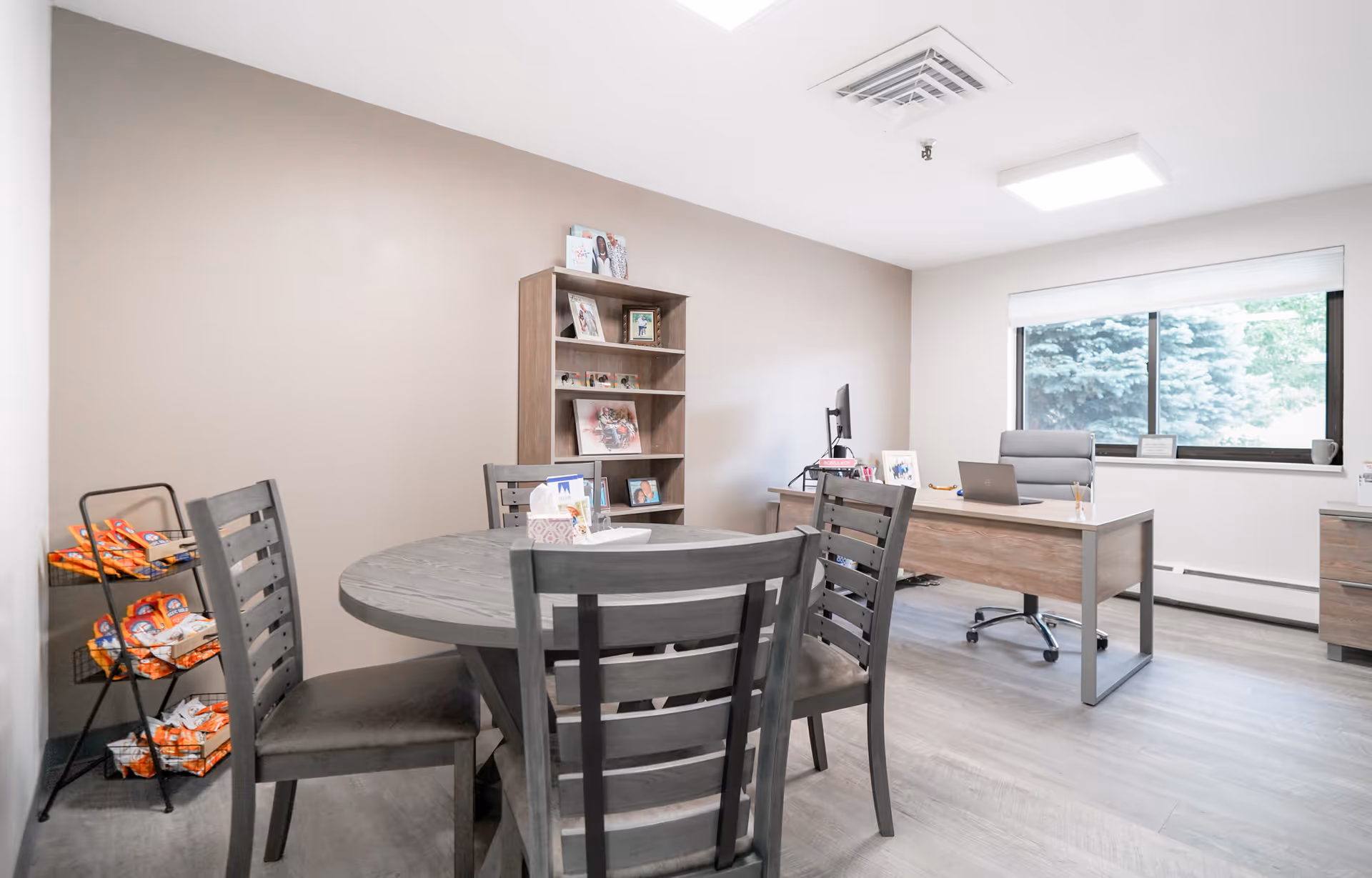 A bright office room with a round wooden table surrounded by four chairs in the foreground. In the background, there is a wooden desk with a computer monitor, laptop, and office chair near a window with a view of trees outside. A wooden bookshelf with framed photos and decorations is against the wall, and a snack rack with various packaged snacks is visible on the left side.