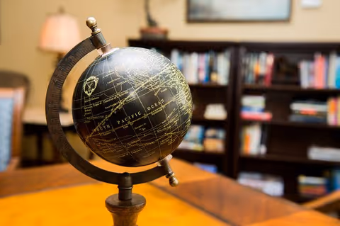 A close-up view of a vintage-style globe on a wooden stand placed on a table, with a blurred background showing bookshelves filled with books and a lamp.
