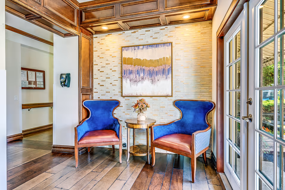 Two blue upholstered chairs flank a small round table with a vase of flowers in a bright seating nook with artwork and glass doors.
